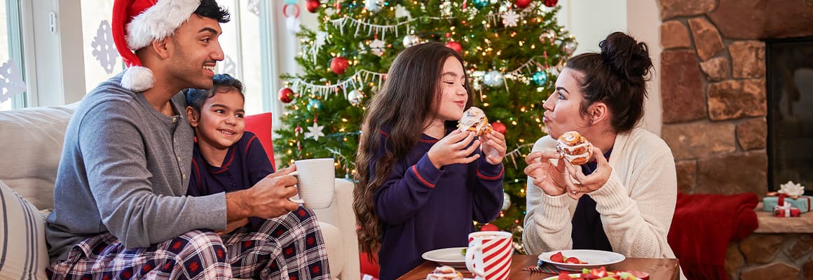 Family wearing pajamas, eating cinnamon rolls and drinking coffee in front of a Christmas tree.