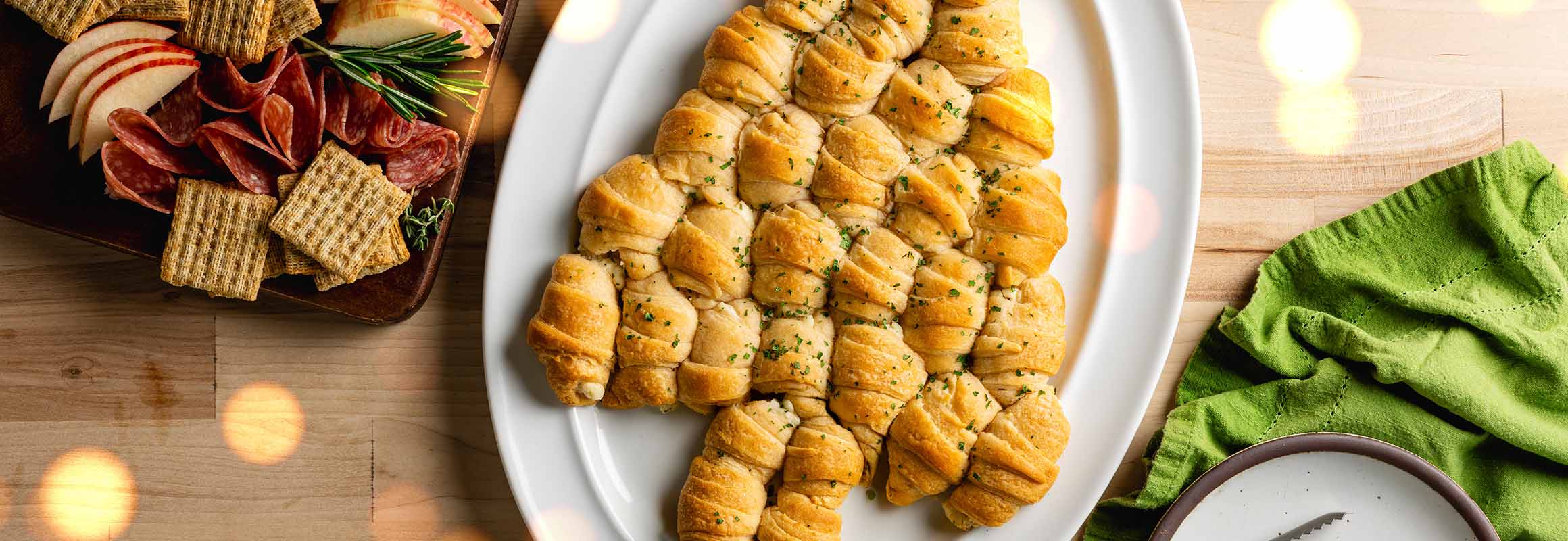 A white serving tray with crescent dough balls filled with cheese that are organized into the shape of a Christmas tree and topped with fresh parsley. A gray and white napkin is in the right hand corner.  