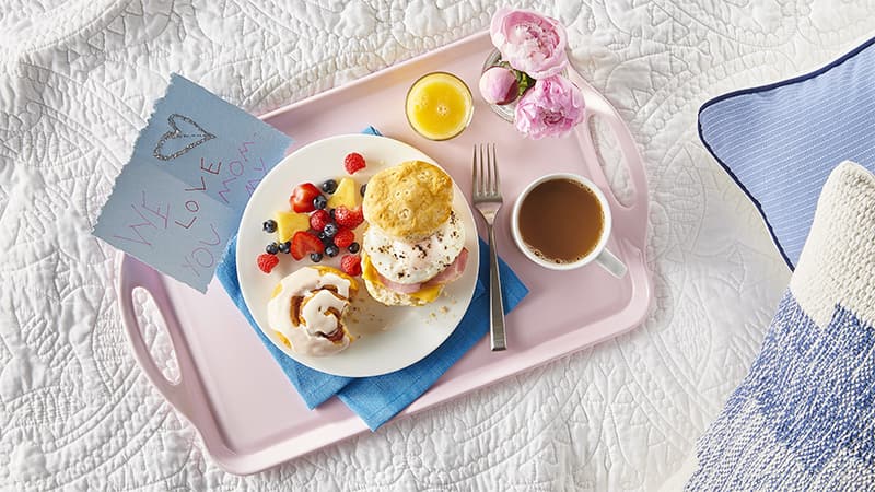 Breakfast tray with a note, orange juice, coffee, flowers, cinnamon roll, fruit, breakfast muffin
