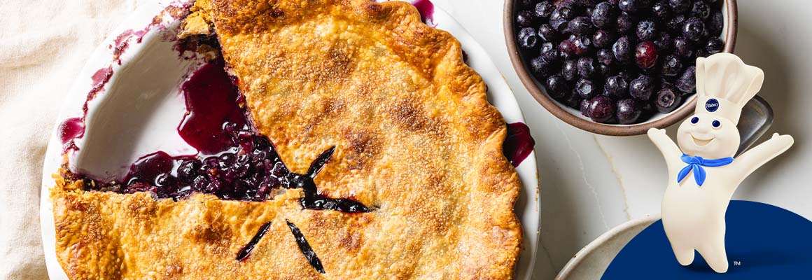 A golden-brown, double-crust blueberry pie baked in a glass dish, featuring a flaky top crust with several steam vents, filled with a bubbling blueberry and tapioca mixture. To the right of the dish there’s a slice of pie on a plate with a fork. In the upper right hand corner of the image there is a bowl of blueberries. 