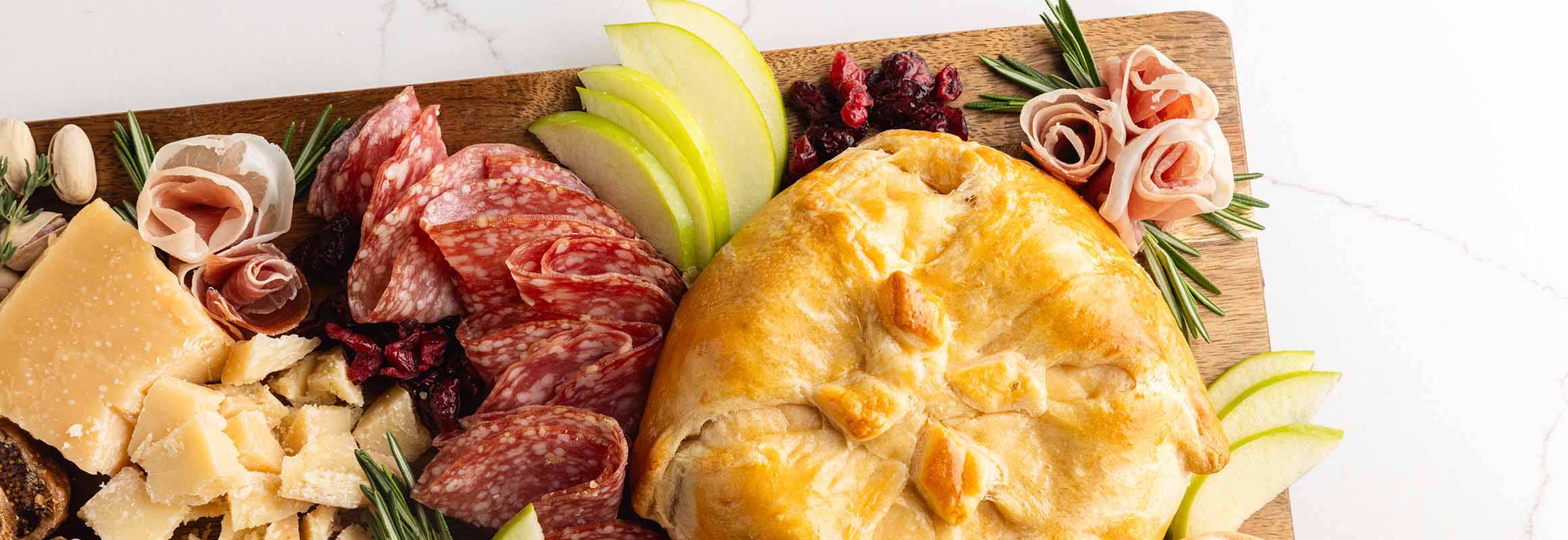 Wooden board serving cheeses, meats, grapes, pears, crackers, and a baked brie.