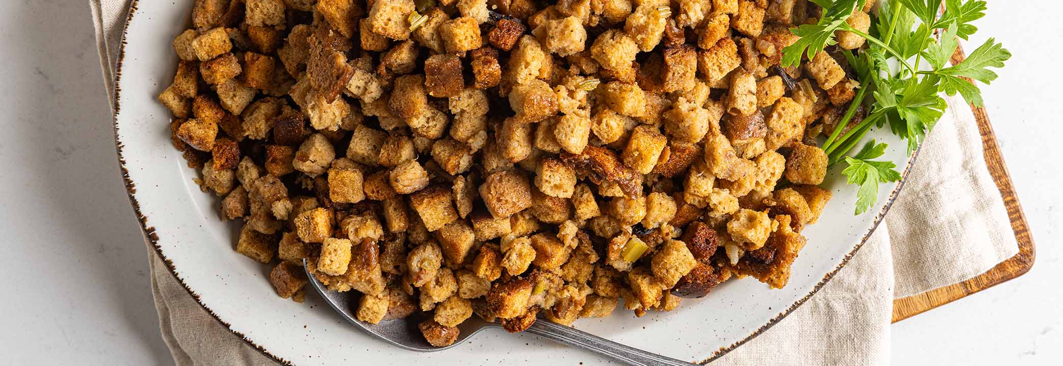 White serving tray with seasoned bread stuffing cubes mixed with celery and onion with parsley garnishing the tray. The tray is placed on top of a cream colored towel and wood cutting board. 