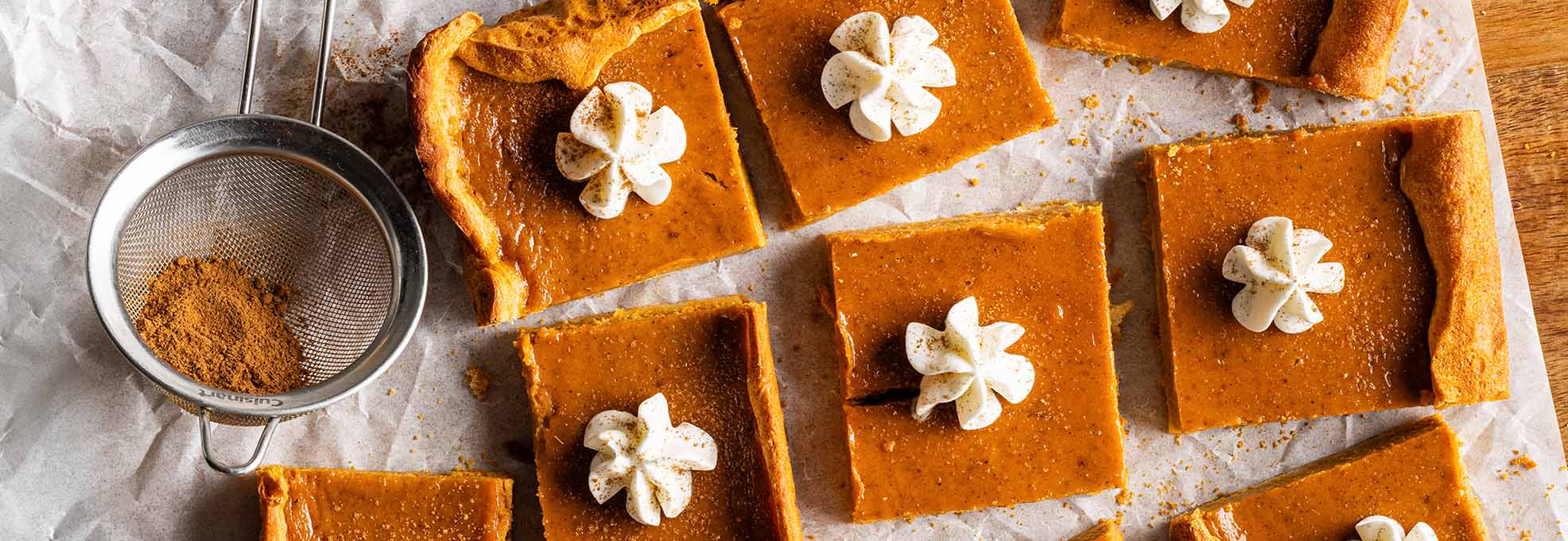 Pumpkin pie bars garnished with whipped cream on top of a sheet of parchment paper on a wood cutting board. 