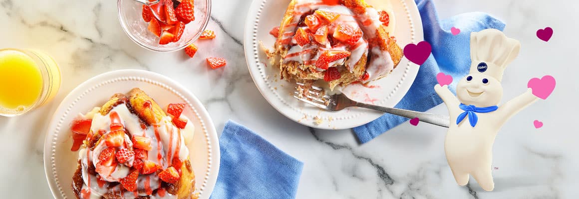 A festive photo of two white plates with baked cinnamon rolls, topped with white icing and chopped fresh strawberries, served on a white marble surface. A glass of orange juice and a small bowl of extra strawberries are also present. The Pillsbury Doughboy character is featured on the right, surrounded by pink and purple hearts.