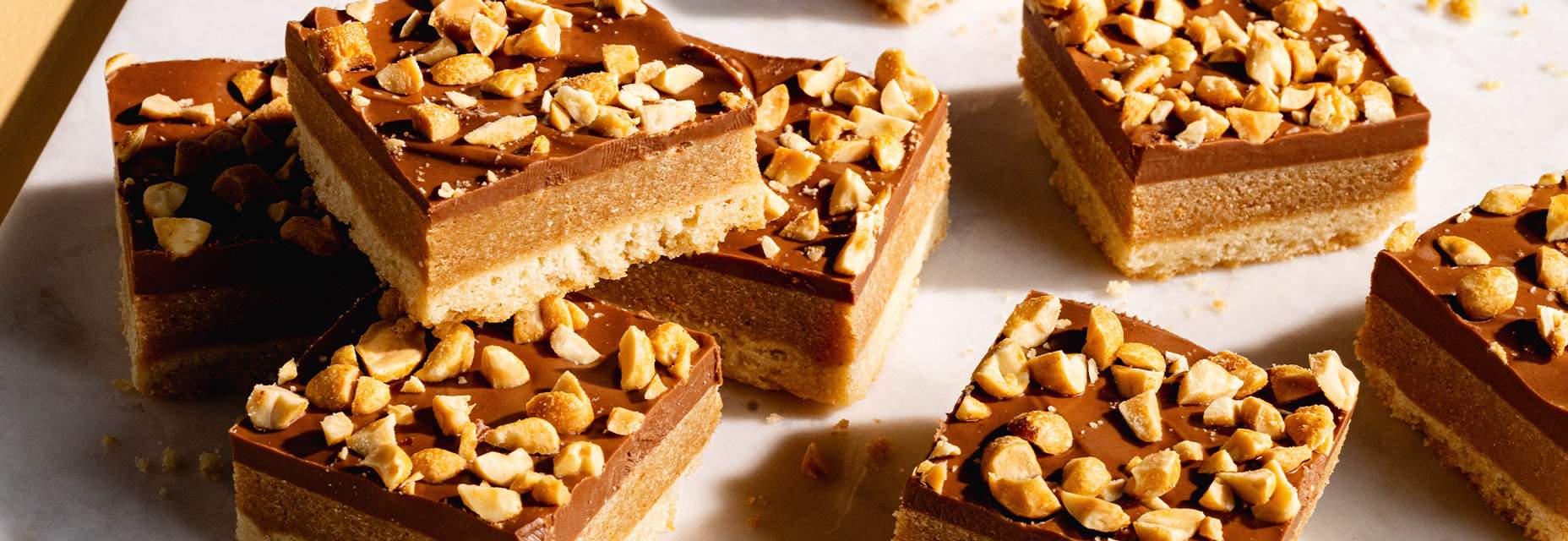 A stack of three-layer dessert bars featuring a golden sugar cookie base, a thick, chewy caramel-graham cracker middle, and a smooth chocolate-peanut butter top layer sprinkled with finely chopped peanuts. 