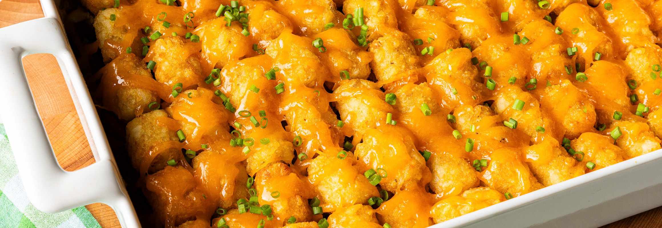 A white casserole dish filled with tater tots, ground beef, cheddar cheese, green beans, and cream of mushroom soup. The cheesy tater tots cover the top of the dish and are garnished with fresh cut herbs. 