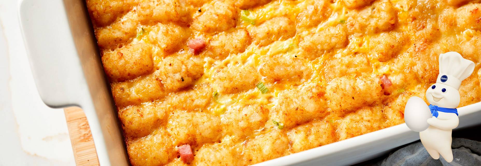 A white casserole dish with an egg bake mixed with cheddar cheese and ham topped with potato puffs. The Pillsbury Doughboy is standing in the far right corner of the image holding an egg.