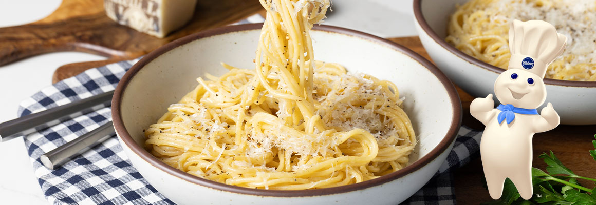 A photo of a white bowl filled with cacio e pepe, a creamy Italian pasta dish topped generously with grated cheese and black pepper. The Pillsbury Doughboy giving two thumbs up in the corner of the image.