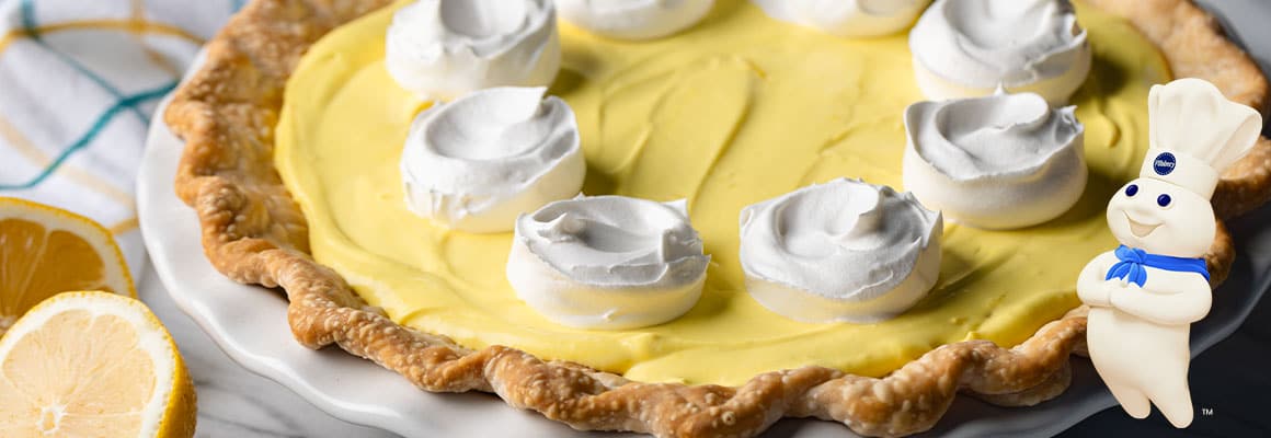 A close-up view of a lemon pie with decorative dollops of white cream or meringue on top, next to half a lemon. The Pillsbury Doughboy in the corner with his arms folded over his stomach.