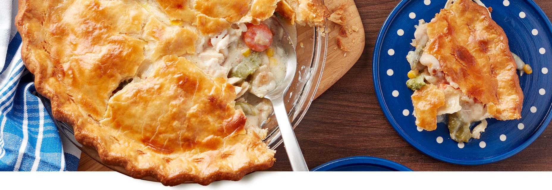 A pot pie with a golden flaky crust with a fluted edge and steam vents exposing a creamy filling with turkey, peas, and carrots in a glass pie plate with a silver spoon for serving. To the right of the plate is a slice of the pot pie on a small blue plate.  