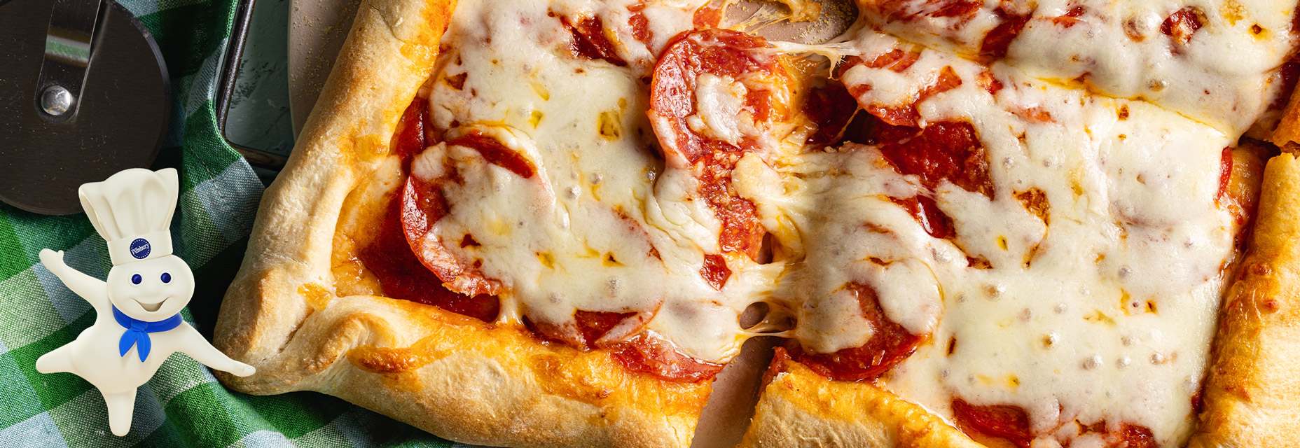 The Pillsbury Doughboy standing on one foot with his arms spread wide in front of an image of a fresh-baked pepperoni pizza with melted mozzarella cheese and a golden-brown crust on a baking stone.