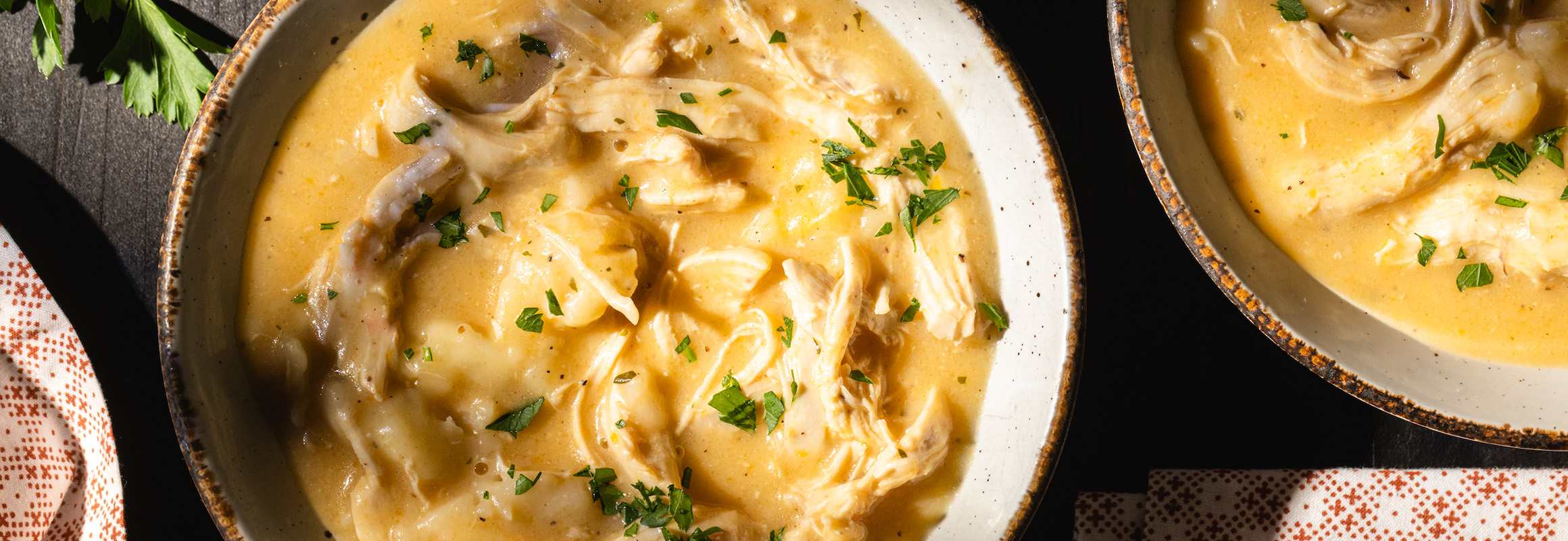 A white bowl of soup filled with chicken broth, shredded cooked chicken, seasonings, biscuits, and cheddar cheese. Topped with fresh herbs. To the right of this bowl is another bowl of soup.
