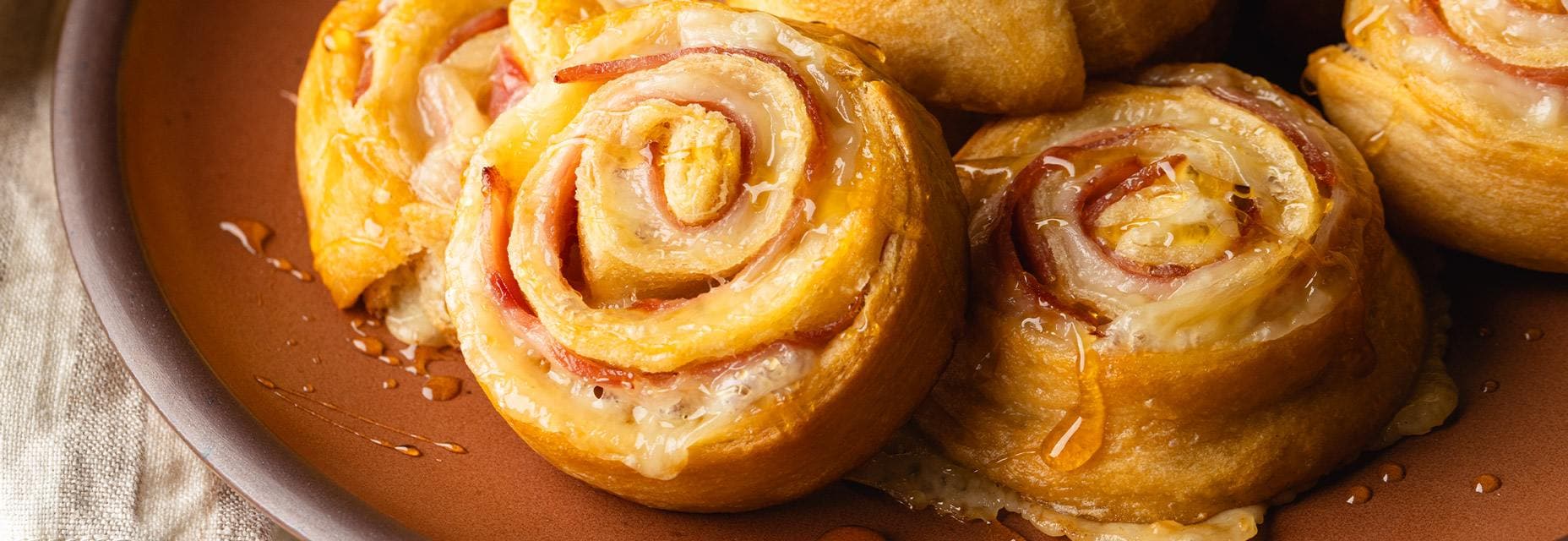 A serving platter holds several golden-baked ham and cheese pinwheels with melted cheese and honey visible.