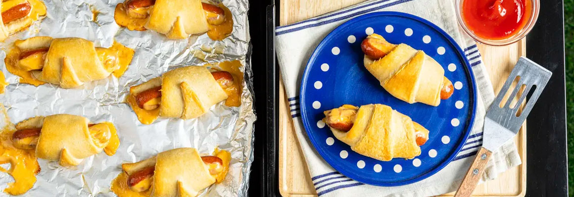 Hot dogs wrapped in crescent rolls being cooked on a grill, served on a blue polka-dot plate.
