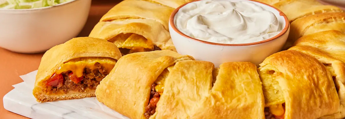 A baked, golden-brown crescent roll ring filled with seasoned beef, nacho cheese-style sauce, tomato, and crushed taco shells. In the middle of the ring there is a bowl of sour cream and in the background there is a bowl of lettuce. 