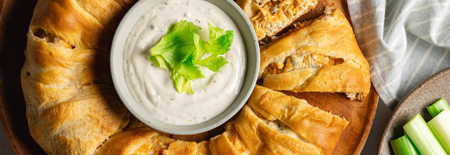 A crescent ring filled with buffalo chicken, with a small bowl of blue cheese dressing in the center and a celery garnish on a wooden serving tray. Next to the tray there’s a plate with celery sticks and a striped towel. 
