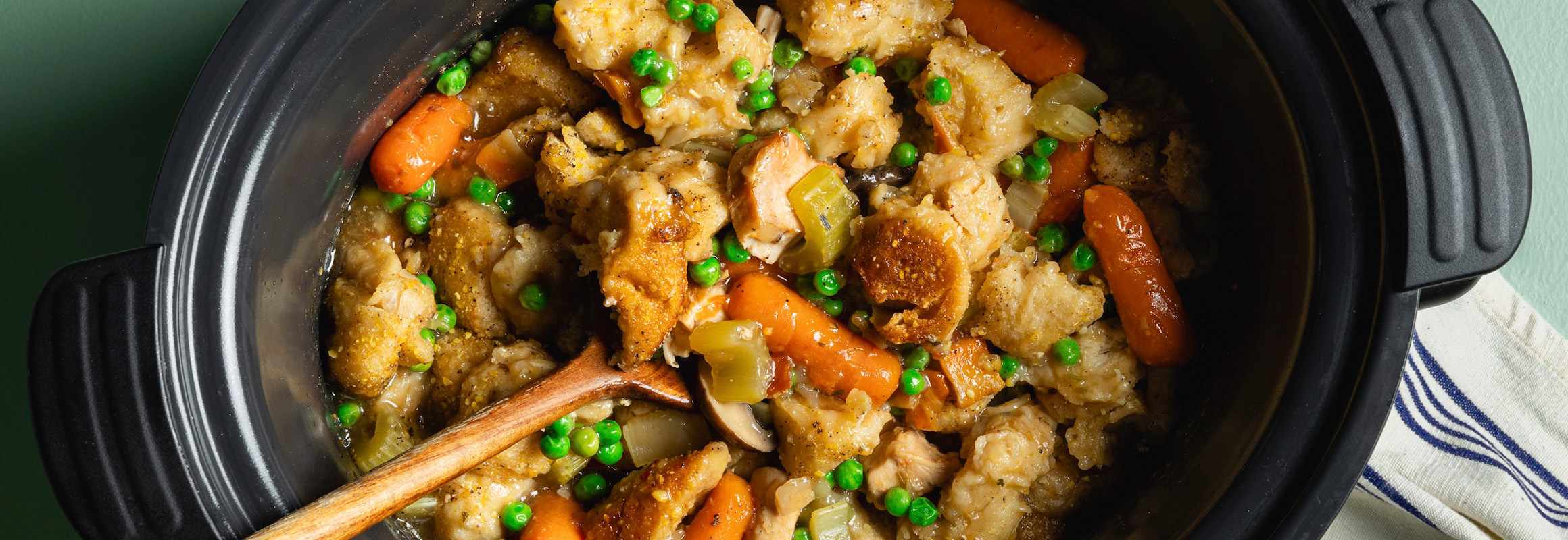 Chicken and dumpling soup in a slow cooker with a wooden spoon inside of it. There are carrots, peas, mushrooms, and celery mixed into the soup. A white and blue towel is to the right of the slow cooker.