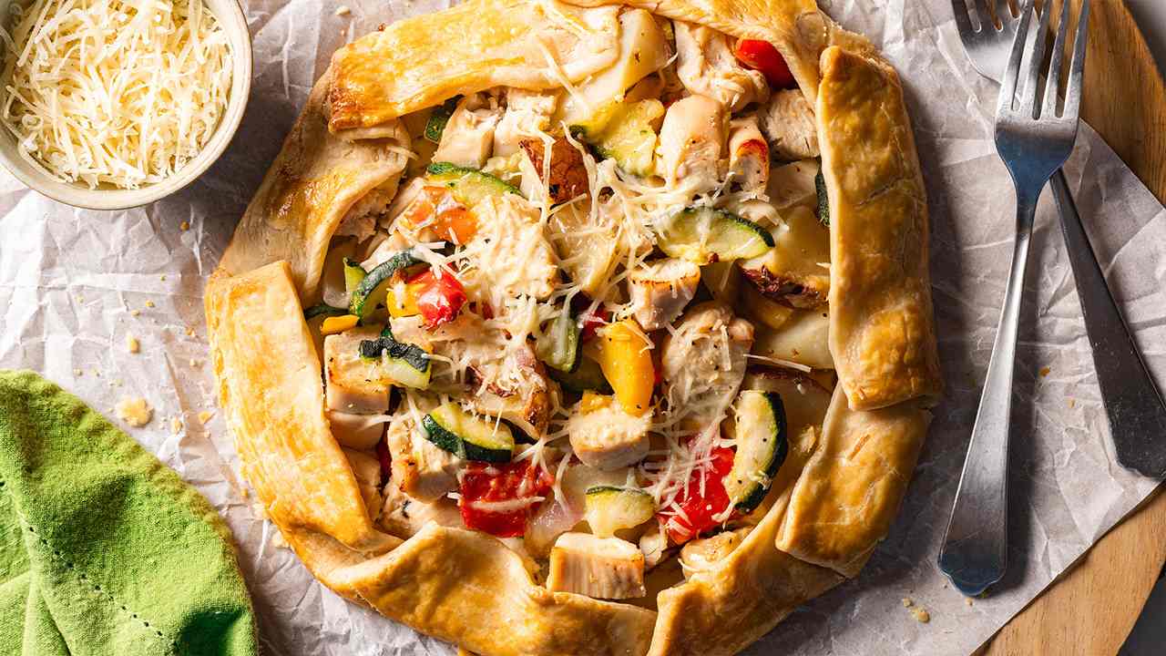 A golden-brown savory galette that has a center filled with a colorful mix of seasoned chicken strips and vibrant Mediterranean-style vegetables, including green beans and red peppers, resting on a creamy bed of ricotta and Parmesan cheese. There’s a bowl of parmesan cheese to the left of the galette and a fork to the right of it. 