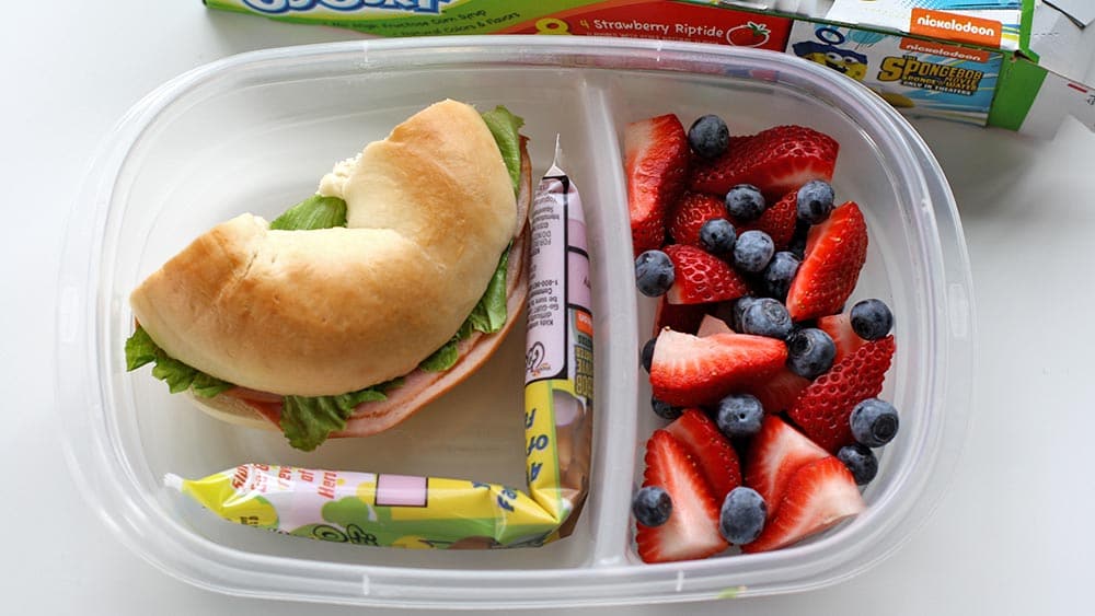 Plastic lunch tray with strawberries, blueberries, half of a bagel sandwich and a Go-Gurt