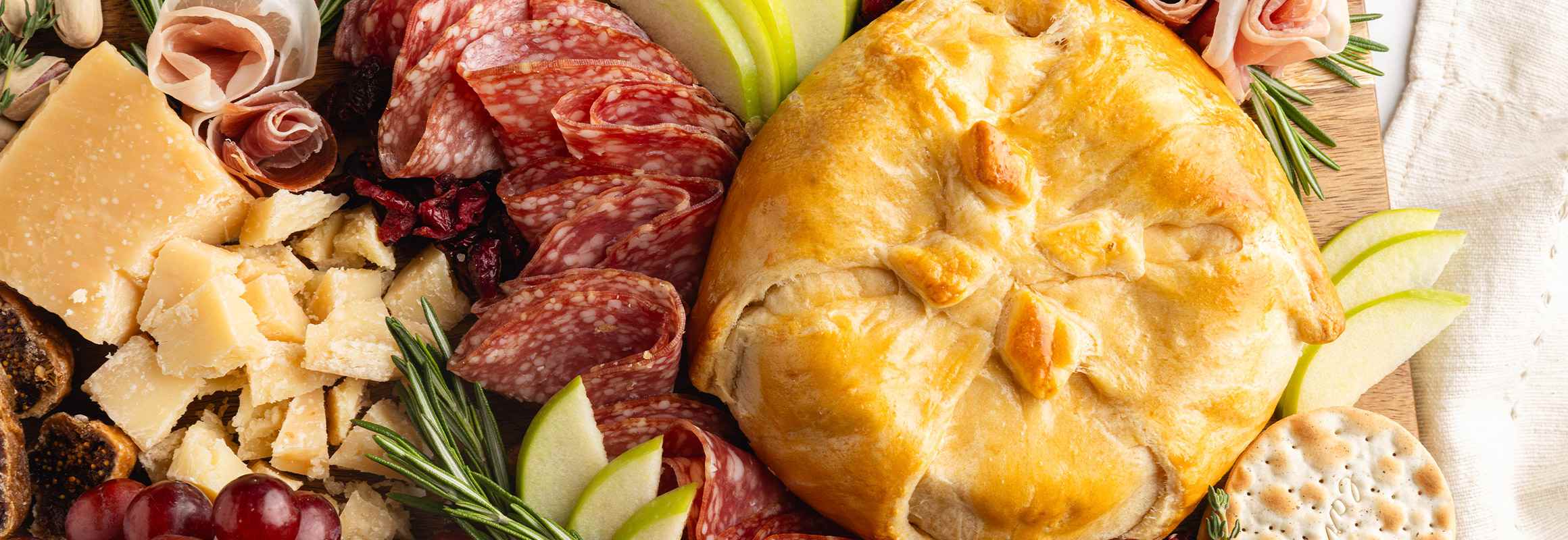 A wheel of warm, golden-baked brie wrapped in flaky crescent dough is presented on a round wooden cutting board, topped with a red jam or preserve and garnished with fresh rosemary sprigs.