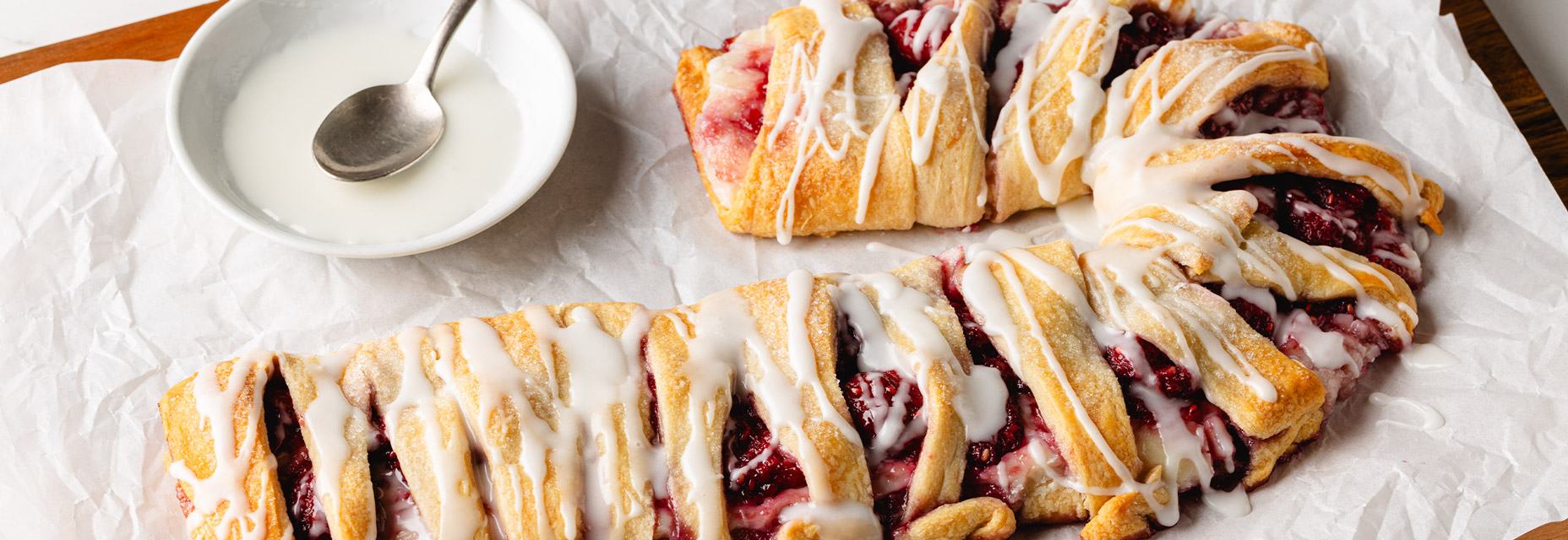 A holiday raspberry cream cheese crescent Danish features a flaky, braided golden crust with red raspberry filling and white icing drizzle, baked in the shape of a candy cane.