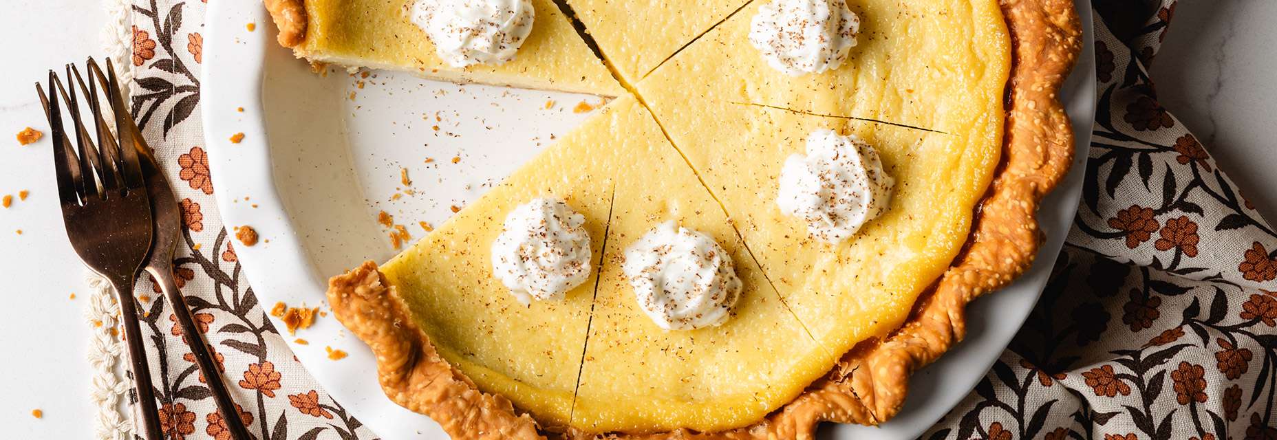 A holiday eggnog custard pie features a golden-baked crust and creamy yellow filling, topped with whipped cream, cinnamon, and nutmeg. A slice is removed, revealing the interior texture of the pie.