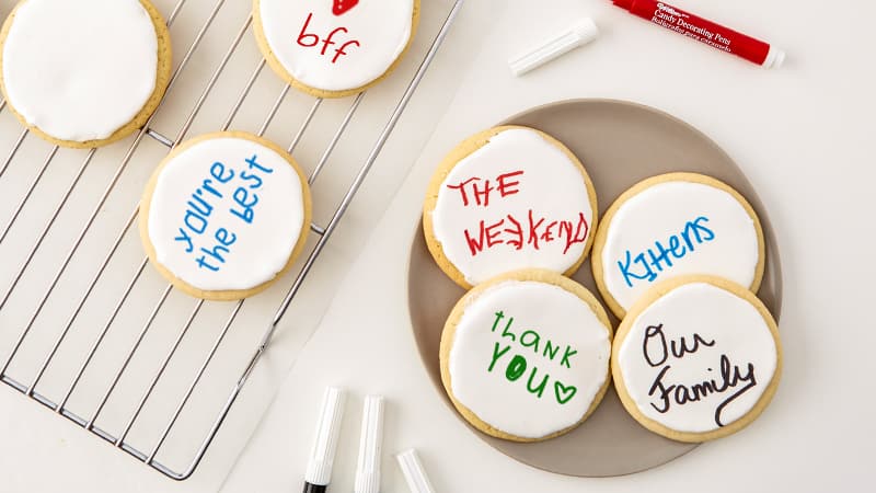 Frosted sugar cookies on a cooling rack and on a plate. Cookies have writing on them. 