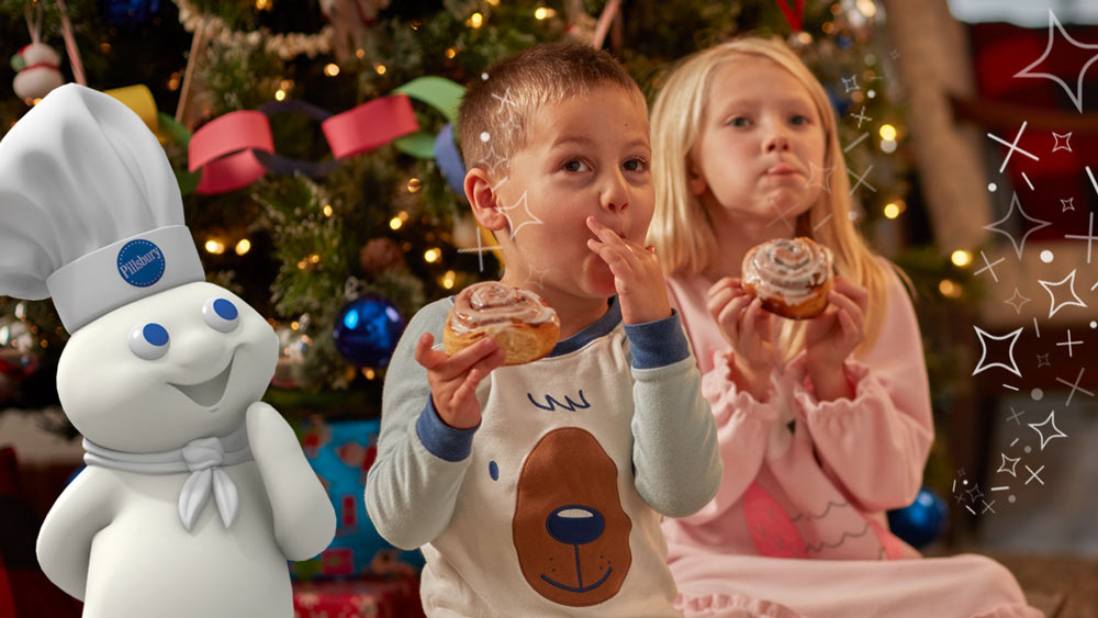 The Pillsbury Doughboy and a boy and a girl eating cinnamon rolls in front of a Christmas tree