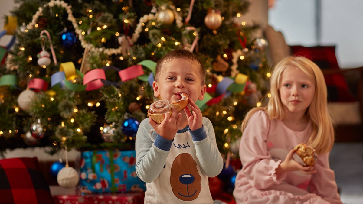 A boy and a girl eating cinnamon rolls in front of a Christmas tree