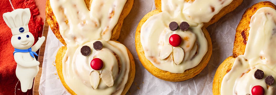 Several baked bunny-shaped cinnamon rolls with white frosting, chocolate chip eyes, and a red candy nose are arranged on a wire cooling rack and a white plate. The Pillsbury Doughboy in the left corner. 