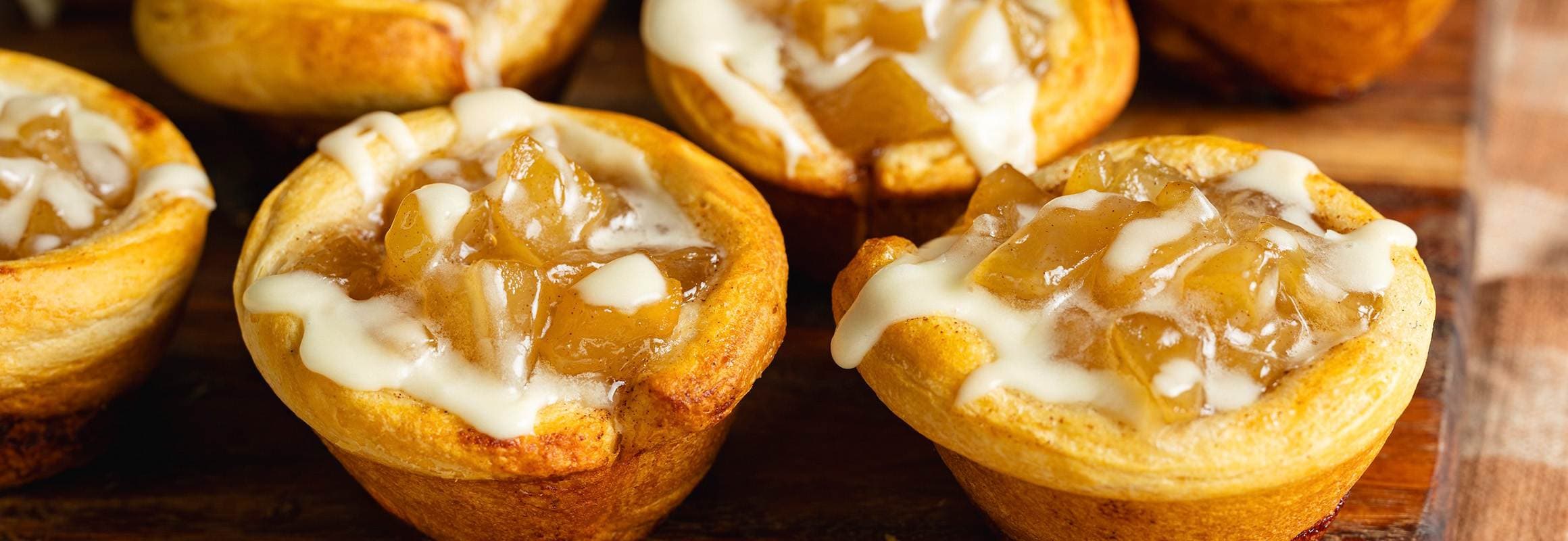 Cinnamon roll cups filled with apple pie filling and cream cheese icing drizzled on top on a wooden board.  