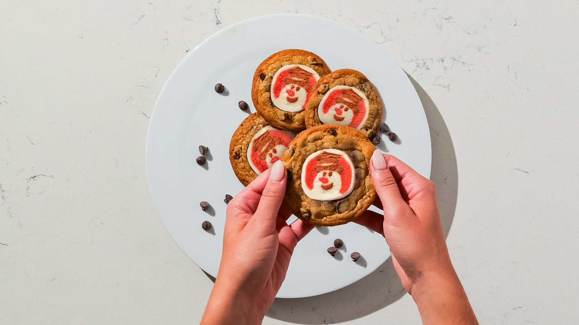 Overhead shot of a person holding a freshly baked chocolate chip cookie topped with a red and white snowman cookie.