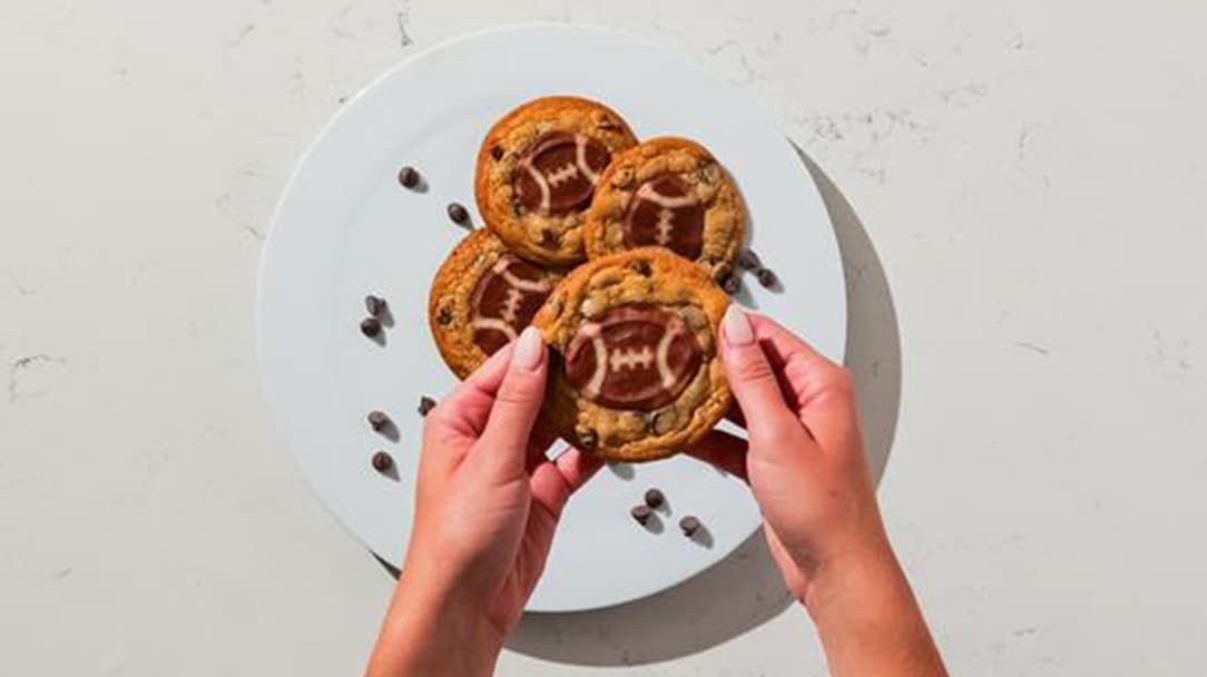 Two hands holding a plate of chocolate chip cookies decorated with football designs, placed on a white marble surface with scattered chocolate chips.