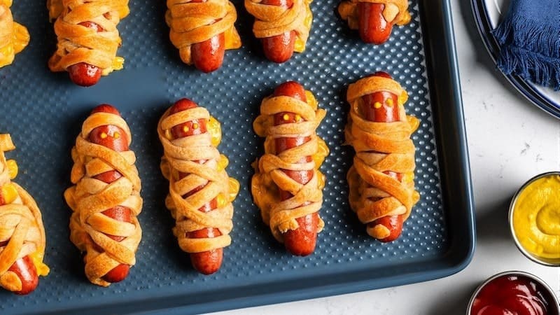 Hot dogs wrapped in dough like mummies with mustard eyes on a baking tray, condiments beside.