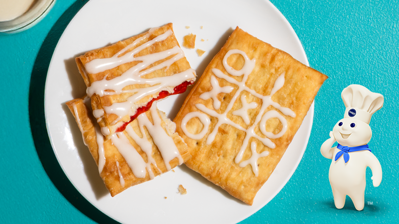Frosted toaster pastries on a white plate, one cut open with red filling, on a teal background with a small dough mascot and a glass of milk nearby.