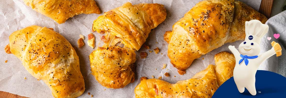 A close-up photograph of baked crescent rolls filled with scrambled eggs and bacon bits. The Pillsbury Doughboy holding a crescent with hearts coming out.