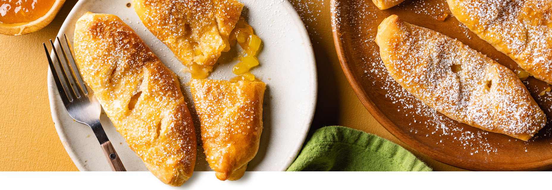A white plate with two flaky pastries filled with sweet jammy pineapple dusted with powdered sugar and a fork. To the right of this plate there are more of the pastries on a wooden plate. A green napkin is in the image as well.