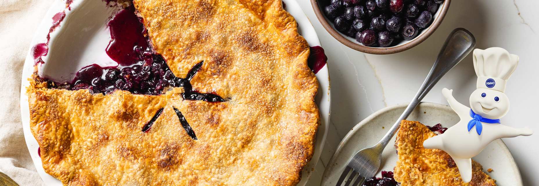 A golden-brown, double-crust blueberry pie baked in a glass dish, featuring a flaky top crust with several steam vents, filled with a bubbling blueberry and tapioca mixture. To the right of the dish there’s a slice of pie on a plate with a fork. In the upper right hand corner of the image there is a bowl of blueberries.