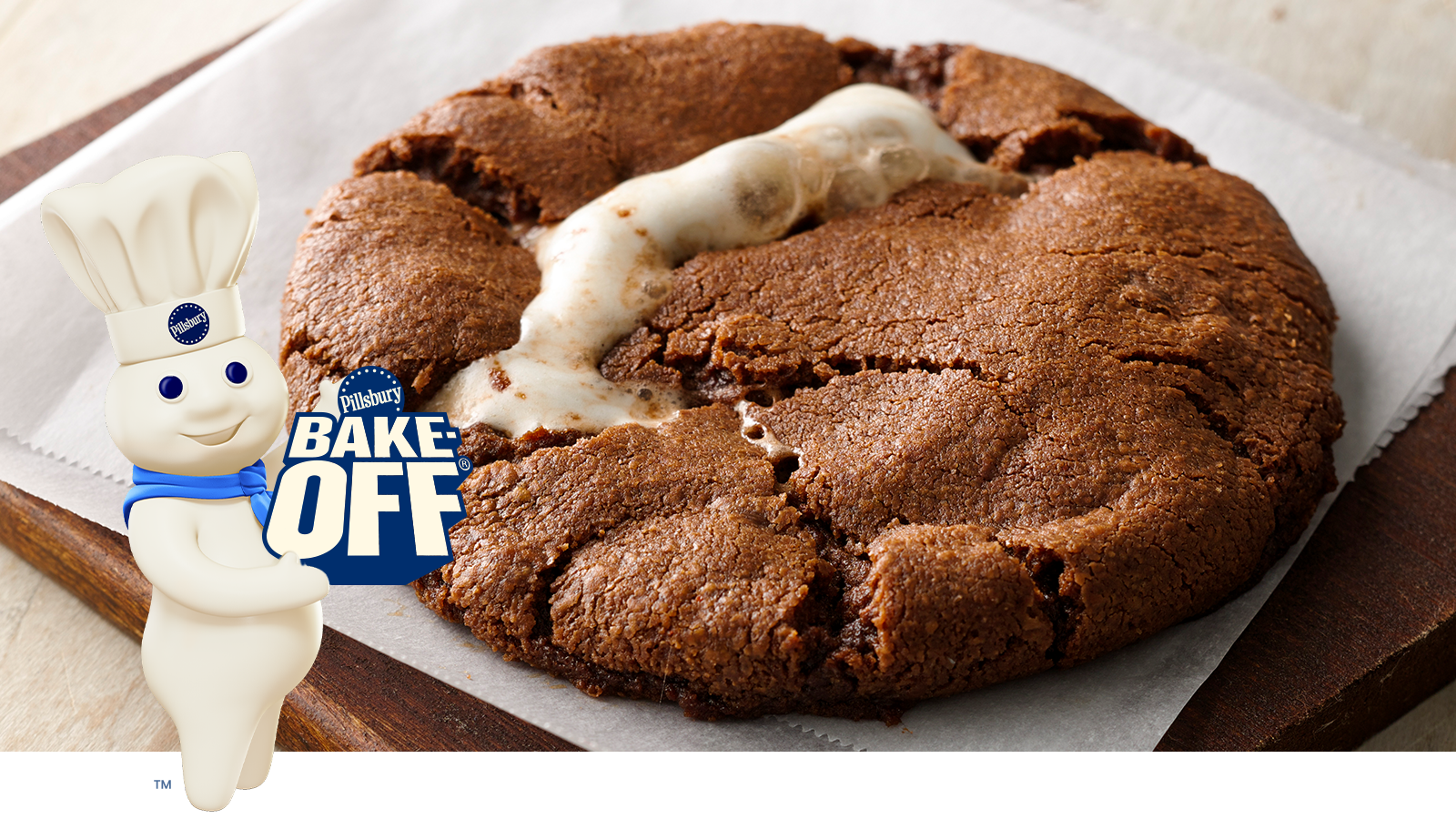The Doughboy holding a sign saying Pillsbury Bake-Off®  in front of a chocolate chip cookie mixed with hazelnut spread, baking cocoa, chili powder, and cinnamon with a marshmallow in the center.