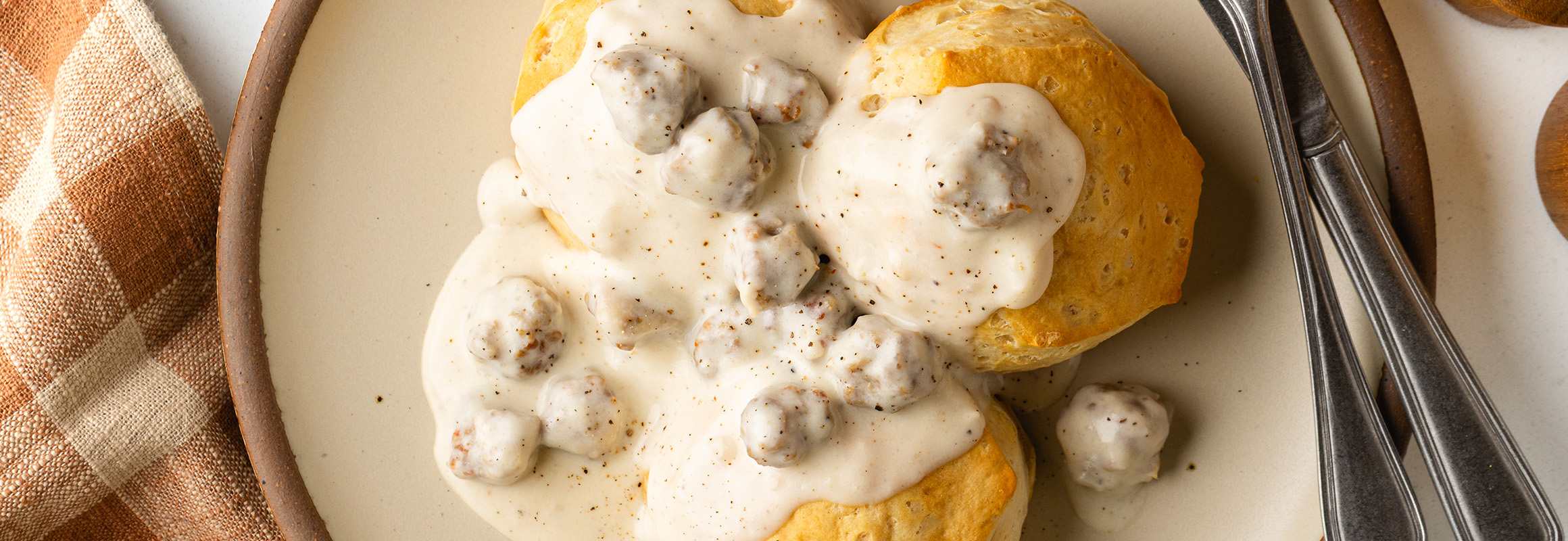 A plate with biscuits covered is sausage and gravy along with silverware. A plaid orange napkin is to the left of the plate.